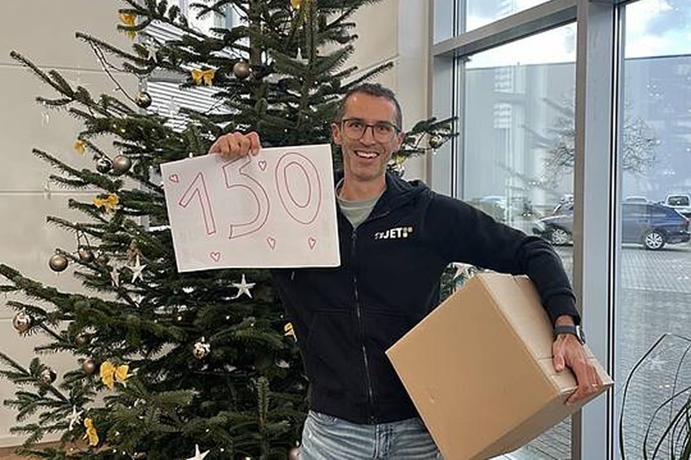 Ein Mitarbeiter von 4JET steht vor einem Weihnachtsbaum. Er hält eine Schachtel und ein Schild mit der Aufschrift „150“ in der Hand.