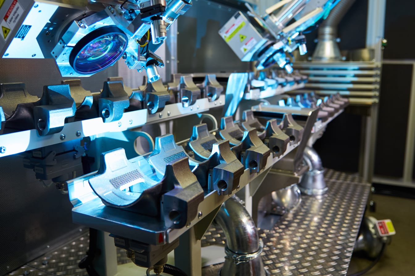 Ein Laserprozesskopf strukturiert ein metallisches Bauteil. Ein heller, blauer Lichtschein ist zu sehen.