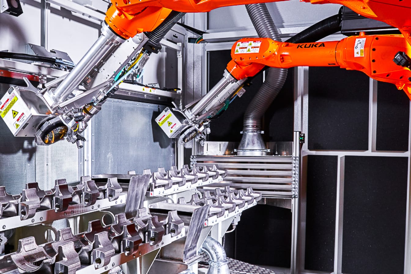 Das Bild zeigt Bauteile, die auf Bearbeitungstischen angeordnet sind und zwei Roboterarme mit jeweils einem Laserprozesskopf, die eine Struktur in die Bauteile einbringen.