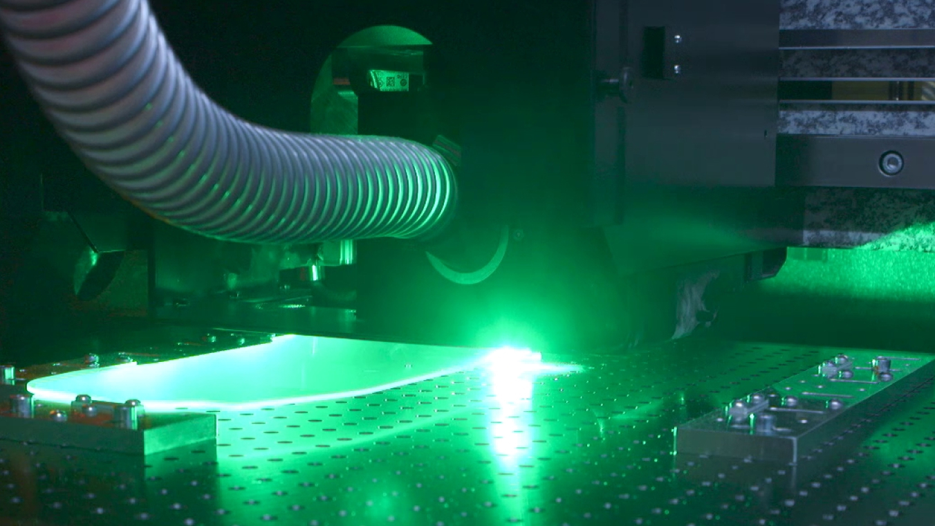 Laserbearbeitungsanlage für Glasbauteile. Das Bild zeigt einen laufenden Prozess mit grünem Leuchten auf einem Freiformbauteil.