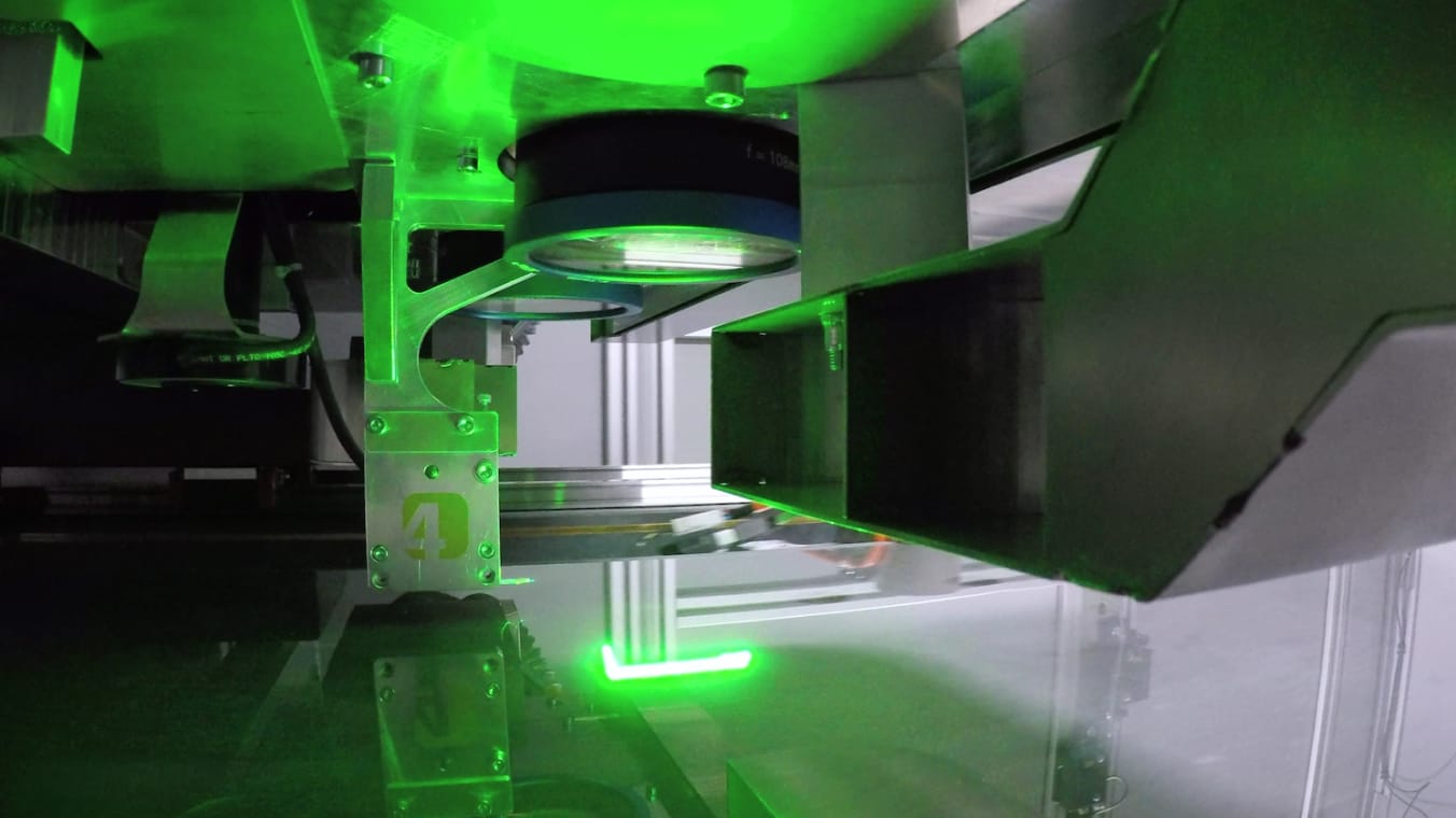 Laserbearbeitungsanlage mit sichtbarem Scanner. Unterhalb des Scanners ist eine Glasscheibe erkennbar. Die Anlage ist grün erleuchtet.