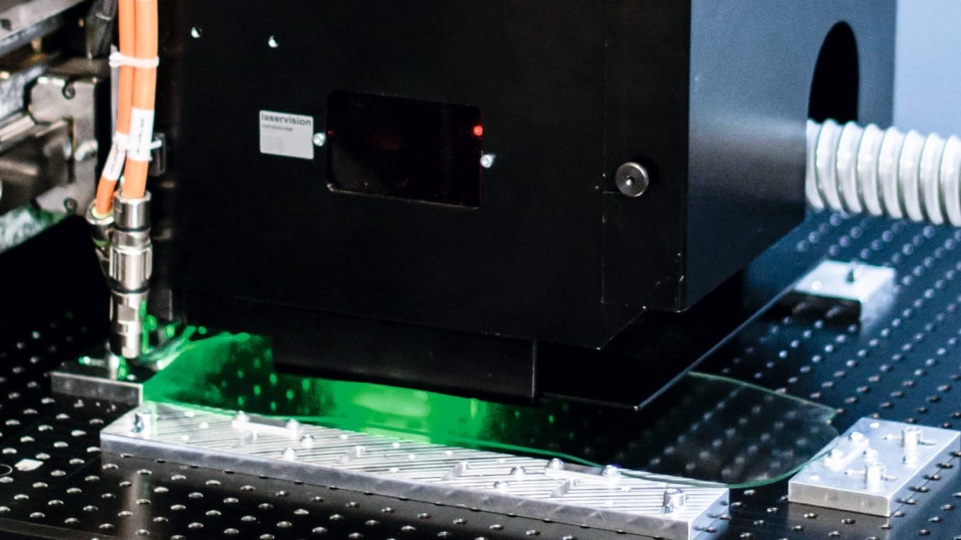 Laserbearbeitungsanlage mit schwarz umhausten Prozesskopf. Unterhalb des Prozesskopfs ist ein grün erleuchtetes Glasbauteil erkennbar.