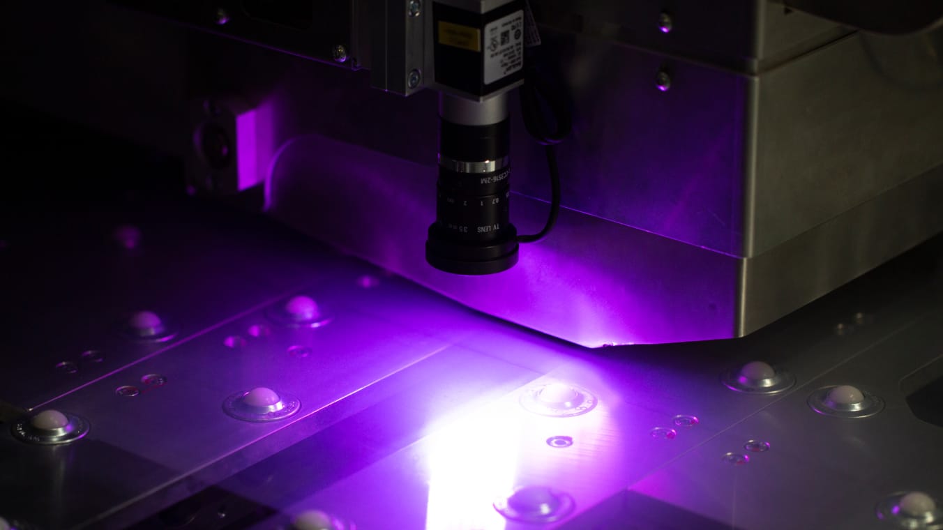 Nahansicht eines Bearbeitungskopfs einer Laseranlage. Unterhalb ist eine blaue erleuchtete Glasscheibe erkennbar.